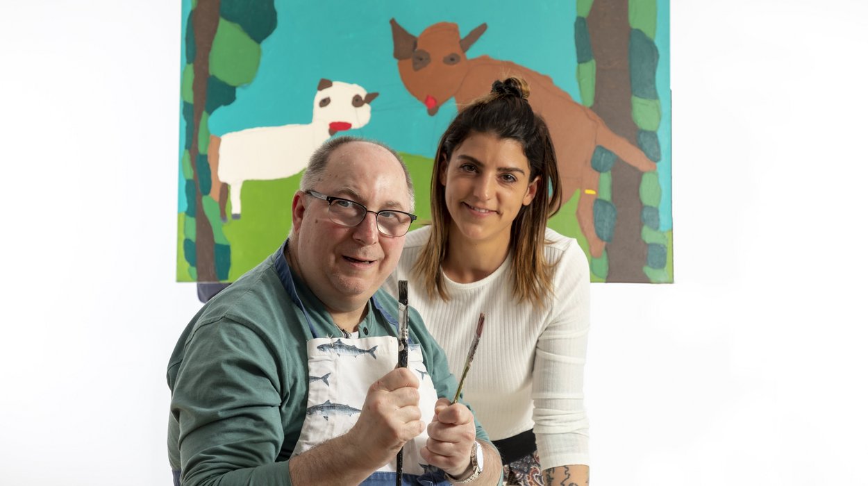 Künstler mit Behinderung und eine junge Mitarbeiterin vor einem Kunstwerk.