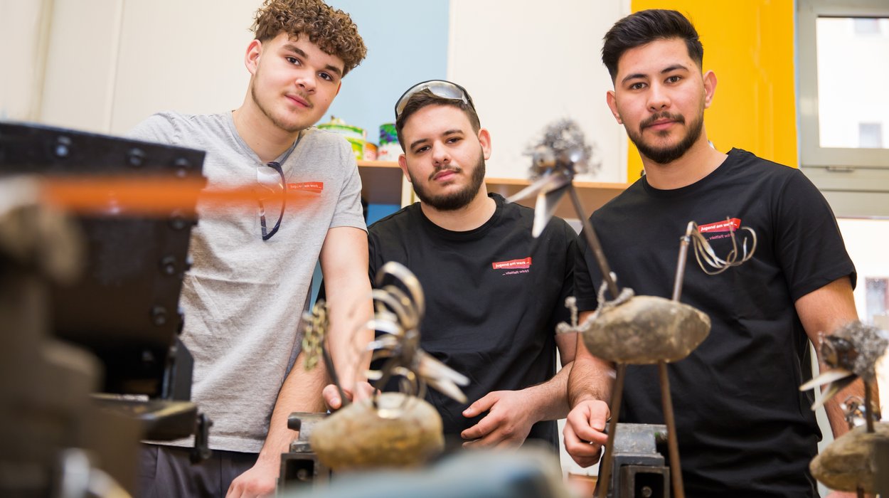Lehrlingscoaching Lehre statt Leere bei Jugend am Werk Steiermark drei Jugendliche blicken in die Kamera, vor ihnen stehen handgefertigte Vögel aus Stahl und Stein