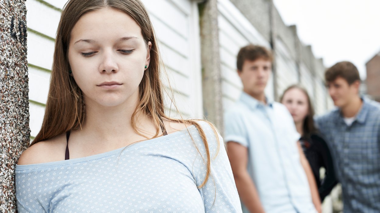 eine Jugendliche mit einem nachdenklichen Blick steht an einer Wand, im Hintergrund zwei Meter weiter schauen drei weitere Jugendliche sie besorgt an