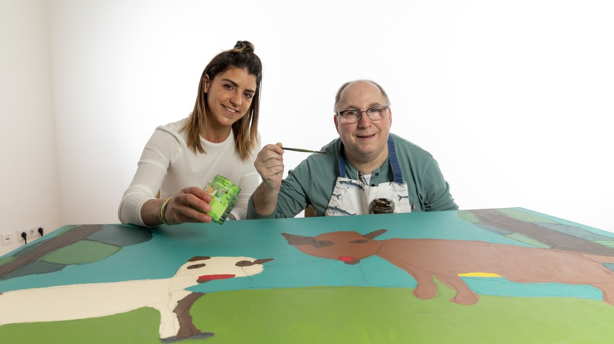 Malwerkstatt ist Atelier und Galerie von Jugend am Werk Steiermark Künstler mit Behinderung und Begleiterin von Jugend am Werk Steiermark sitzen mit Pinseln und Farbe bei einem Bild