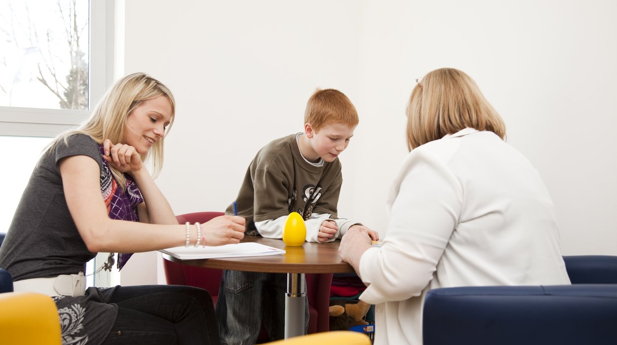zwei Damen und ein Kind sitzen bei einem Tisch, die eine schreibt auf einem Block mit und die andere spielt mit dem Kind
