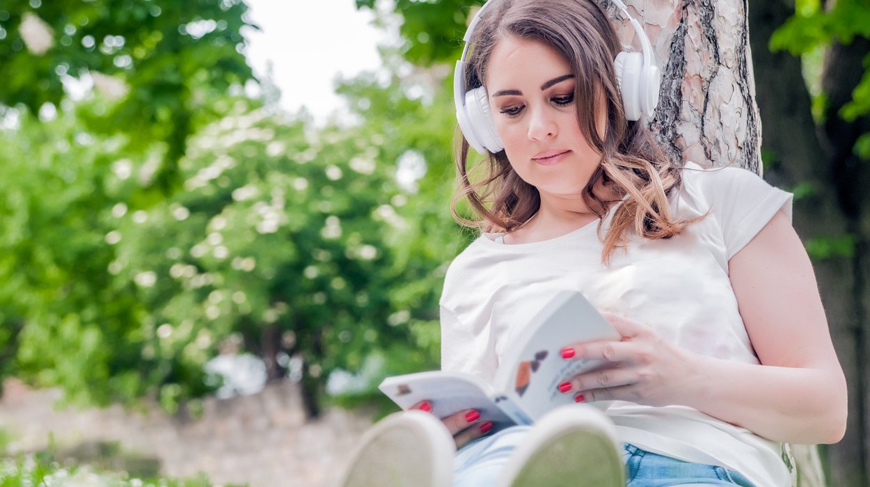 Eine Frau sitzt an einen Baum gelehnt im Park und liest in einem Buch, sie trägt Kopfhörer