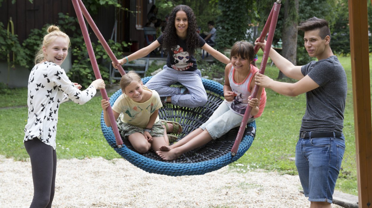 drei Kinder sitzen in einer Korbschaukel während zwei Betreuer*innen sie antauchen