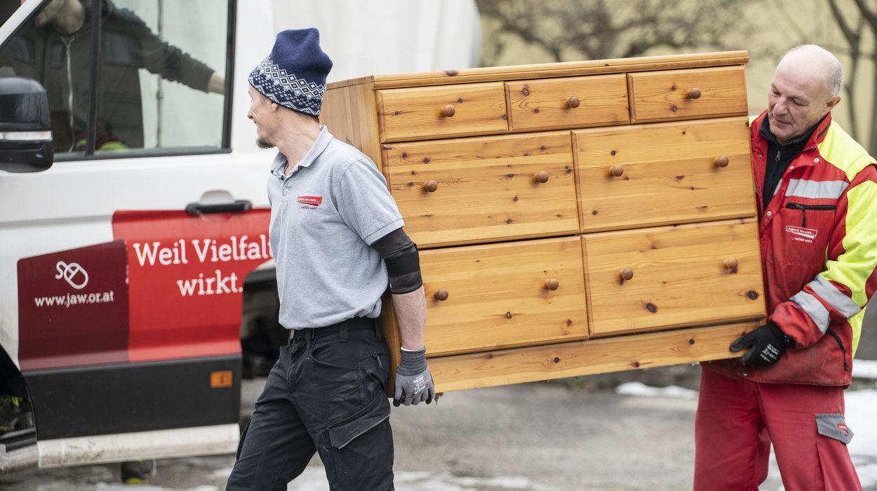 Enturempelung_Jugend-am-Werk-Steiermark Zwei Männer tragen eine Holzkommode zu einem Fahrzeug von Jugend am Werk.