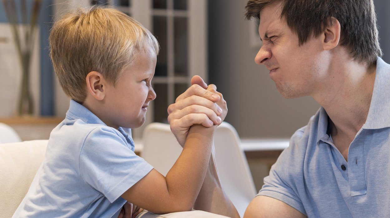 ein kleiner Junge macht Armdrücken mit einem Mann, beide schauen sich mit drückenden Gesichtsausdrücken an