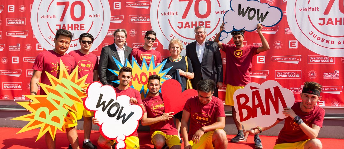 Zwei Männer im Anzug sowie eine Gruppe Jugendlicher in roten T-Shirts steht vor einer Wand mit der Aufschrift "70 Jahre Jugend am Werk", sie alle halten Schilder mit Aufschriften wie "Bamm" und "Wow" hoch