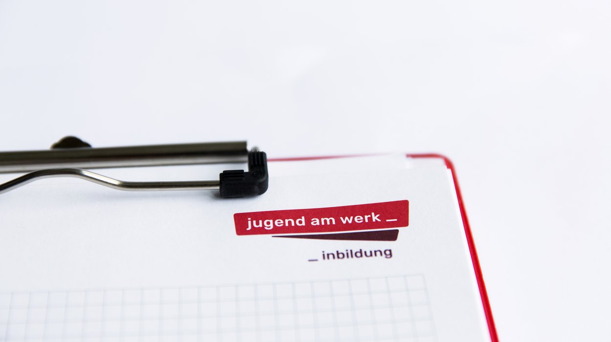 inbildung_Jugend-am-Werk-Steiermark Ein Klemmbrett mit einem karierten Zettel darauf, in der rechten Ecke ist das Logo von Jugend am Werk Steiermark zu sehen