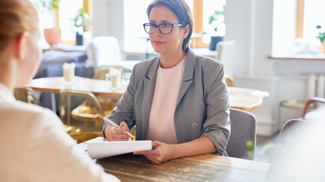 Service_Jugend-am-Werk-Steiermark Eine Frau sitzt an einem Tisch und bespricht etwas mit einer anderen Frau, die mit dem Rücken zum Betrachtenden im Bild sitzt