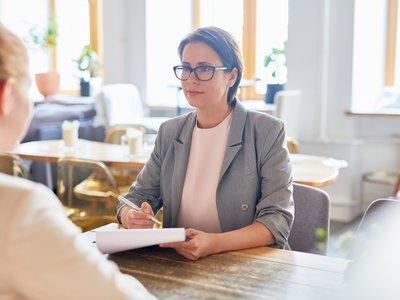 Service_Jugend-am-Werk-Steiermark Eine Frau sitzt an einem Tisch und bespricht etwas mit einer anderen Frau, die mit dem Rücken zum Betrachtenden im Bild sitzt