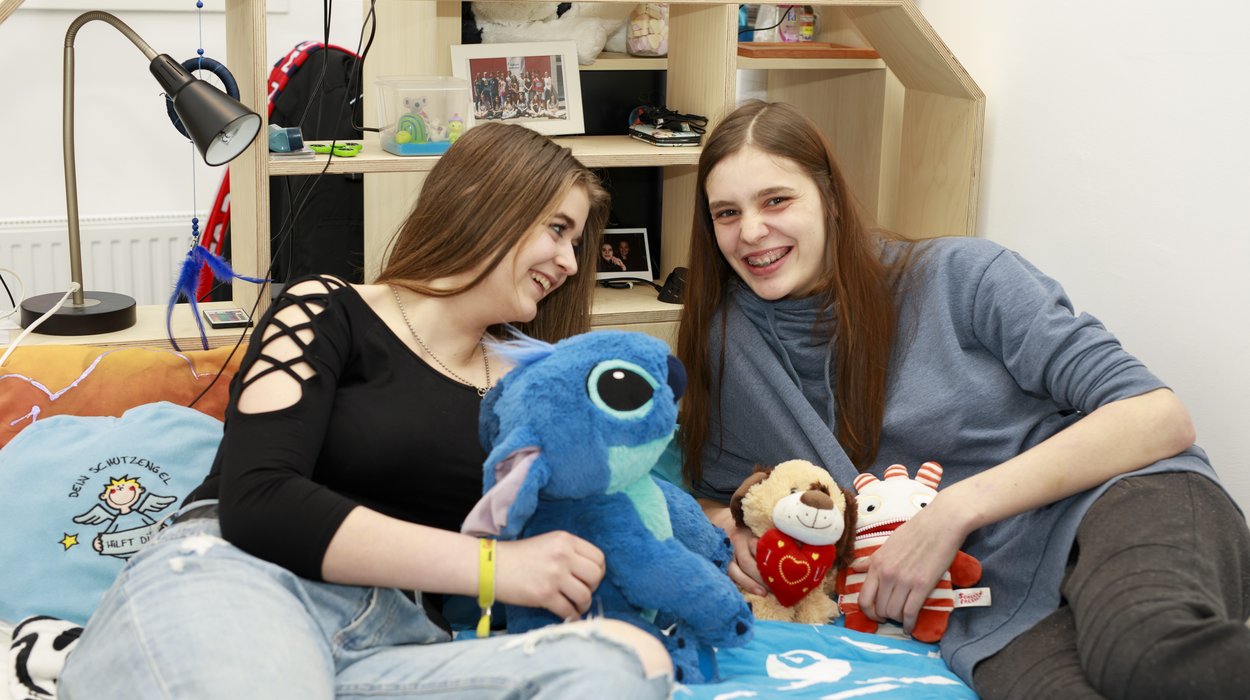 Sozialpaedagogische-Wohngemeinschaft_Jugend-am-Werk-Steiermark zwei Jugendliche sitzen lachend auf einem Bett in einem gemütlichen Raum mit guter Atmosphäre