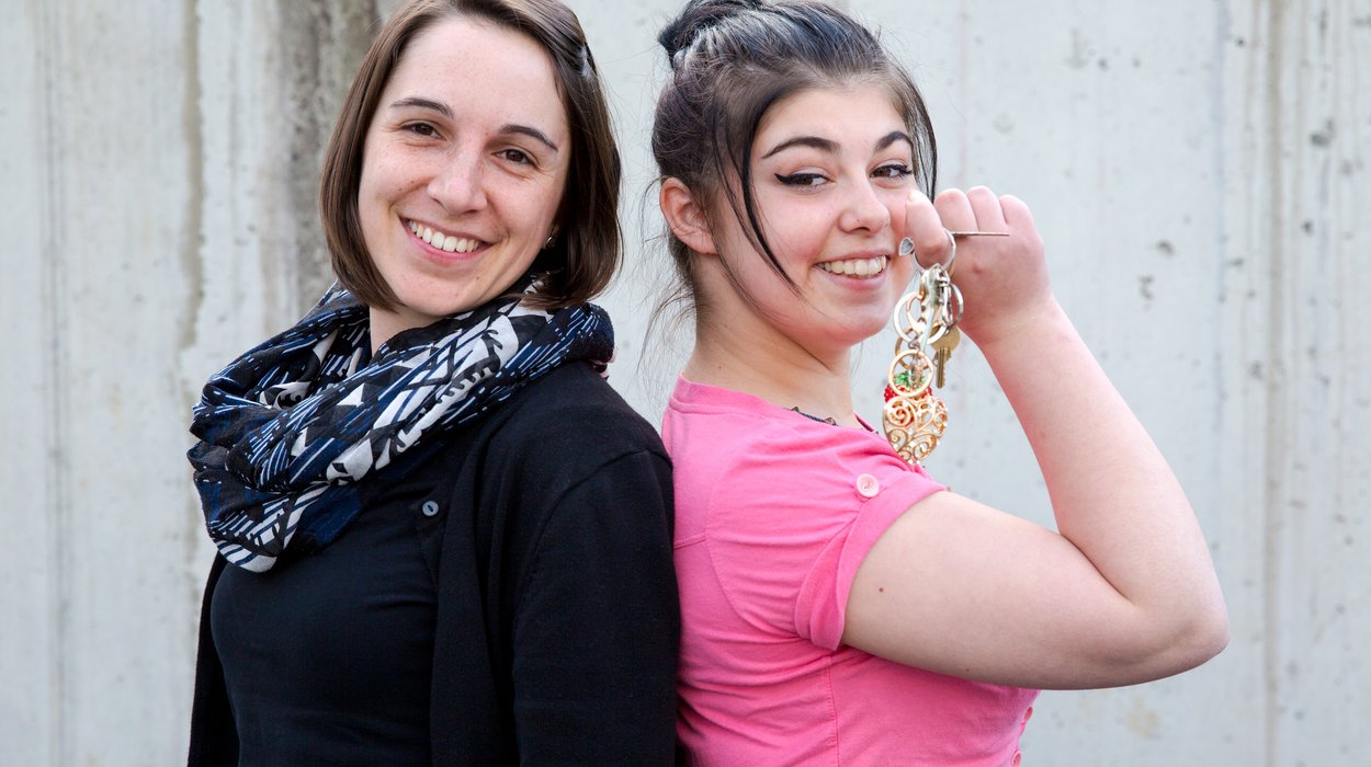 Betreutes-Wohnen_Jugend-am-Werk-Steiermark eine Jugendliche mit einem Schlüssel und eine Betreuerin stehen freudig lachend Rücken an Rücken