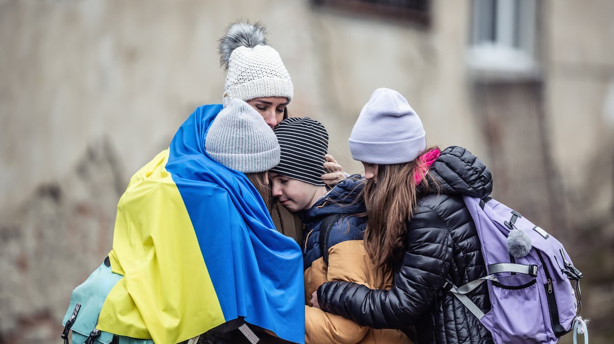 eine Flüchtlingsfamilie aus vier Personen ohne Vater hält sich zusammen umarmend mit einer ukrainischen Flagge warm