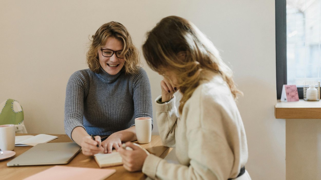 Zwei Frauen sitzen an einem Tisch, die eine erklärt der anderen etwas und zeigt dabei auf ein Blatt Papier zwischen ihnen