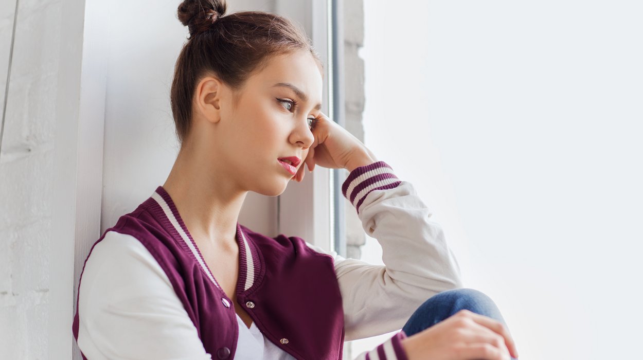 Eine Jugendliche sitzt an einem Fenster, den Kopf mit der Hand aufgestützt und schaut nachdenklich nach draußen