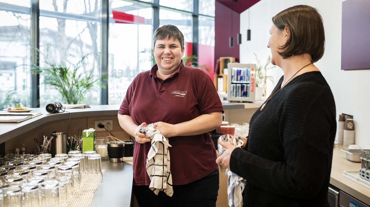 Eine Frau mit Behinderung trocknet Gläser ab, eine Begleiterin unterstützt sie dabei