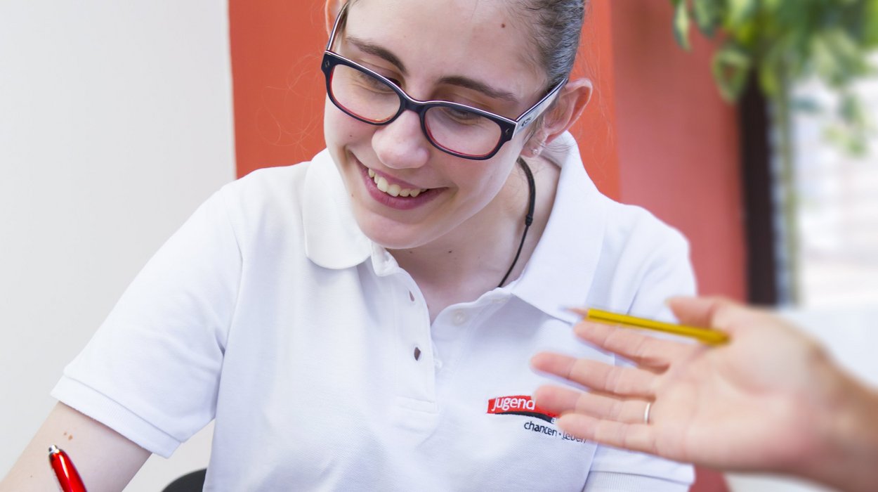 inberatung_Jugend-am-Werk-Steiermark Eine Frau mit Behinderung sitzt an einem Tisch und schreibt etwas, während eine andere Person, die nicht ganz im Bild ist, ihr etwas erklärt
