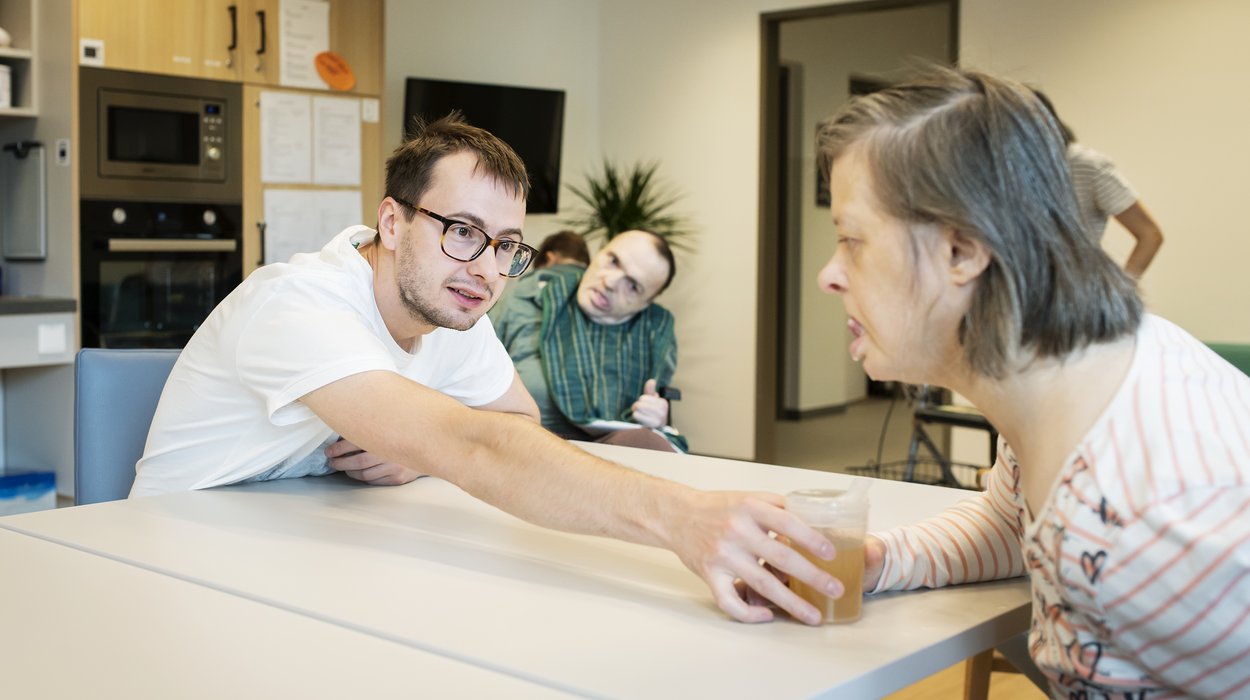 Ein Begleiter reicht einer Frau mit Behinderung ein Glas über den Tisch