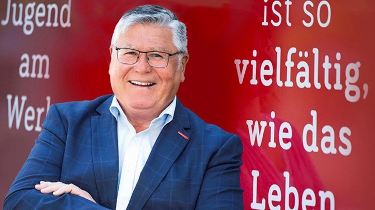 Ein Mann in einem Anzug vor einer roten Wand mit der Aufschrift "So vielfältig wie das Leben"