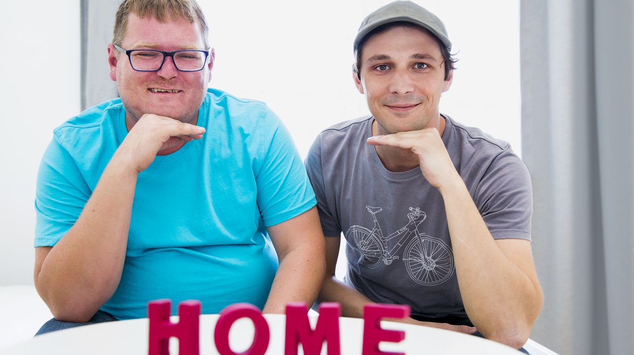 eine Person mit Behinderung sitzt neben einer anderen Person, beide halten ihre Hand unter dem Kinn