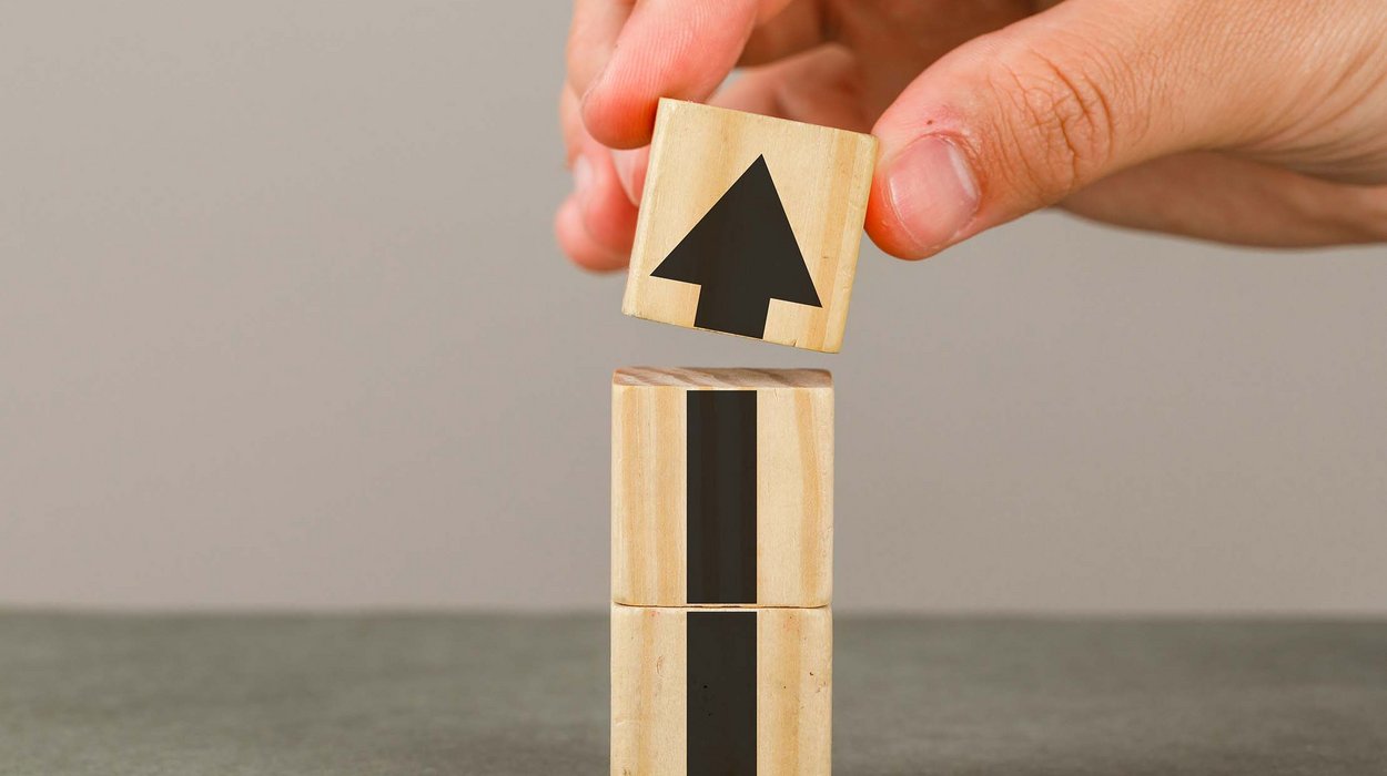 Eine Hand stellt drei Holzwürfel übereinander auf, auf den Holzwürfeln ist ein Pfeil nach oben zu sehen
