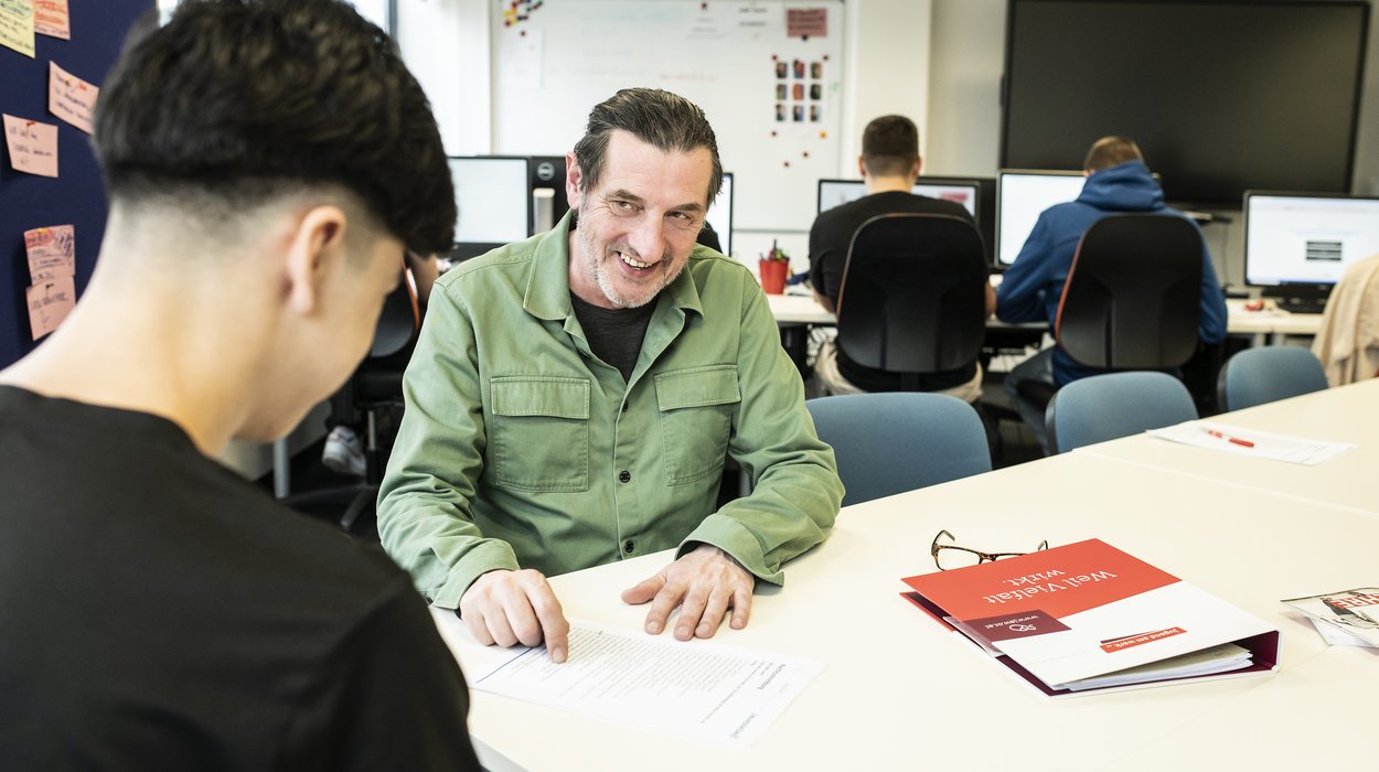 Ein Mann berät einen Jugendlichen in einer Büroräumlichkeit, im Hintergrund sind weitere Jugendliche an PCs zu sehen