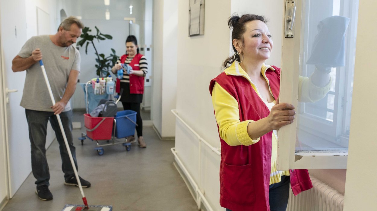Reinigungsservice_Jugend-am-Werk-Steiermark In einem Gang: Frau putzt Fenster, Mann wischt den Boden, weitere Frau steht neben einem Putzwagen.