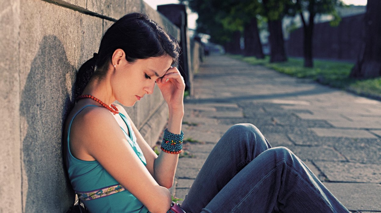 eine Jugendliche sitz mit verzwicktes Bodenschauen und Hand an der Stirn am Boden an einer Wand