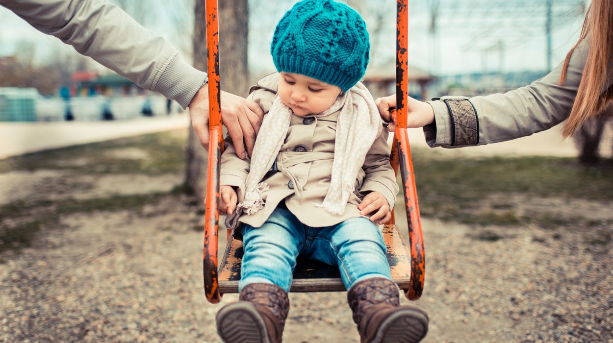 ein kleines Mädchen sitzt auf einer Kinderschaukel, ihre beiden Elternteile schupfen sie gemeinsam an mit jeweils einer Hand