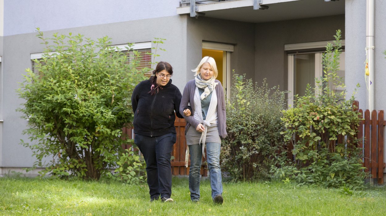 Wohnverbund_Kapfenberg_Jugend-am-Werk-Steiermark eine Person mit Behinderung geht mit einer Betreuerin Arme eingehakt im Garten