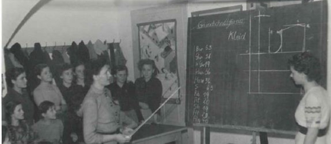 Ein schwarzweißes Foto, zwei Frauen erklären anhand von Zeichnungen auf einer Tafel einer Gruppe Mädchen ein Schnittmuster für ein Kleid