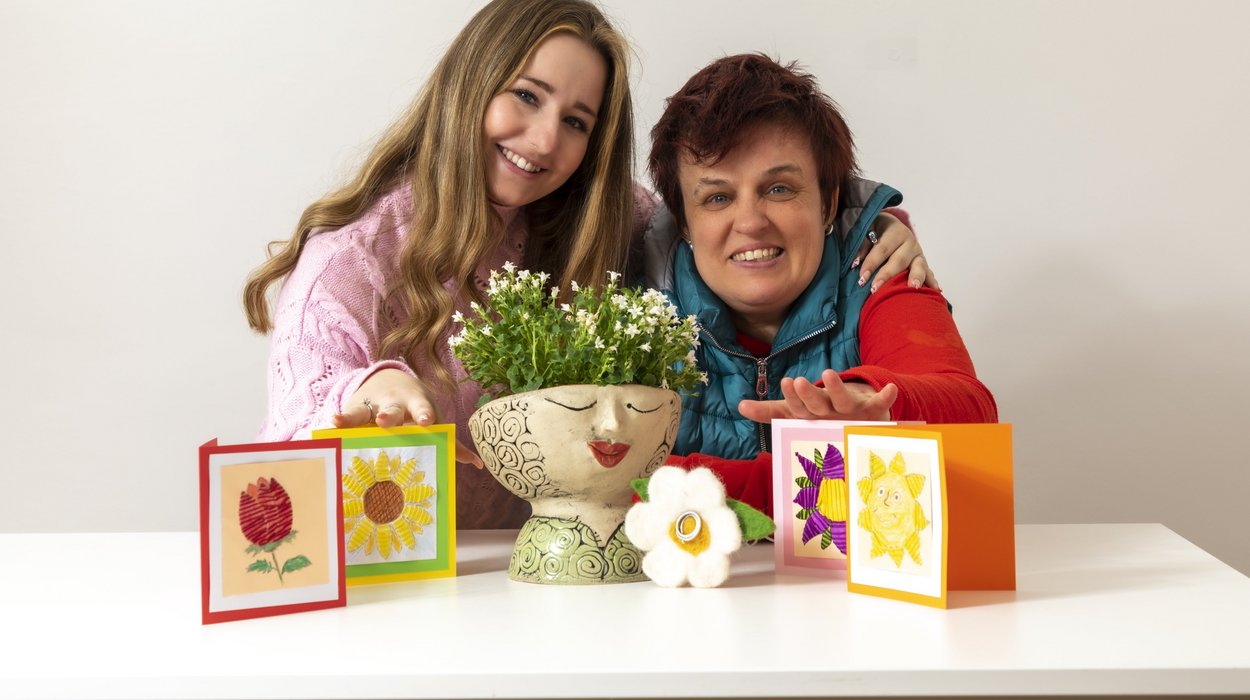 Spenden_Jugend-am-Werk-Steiermark Eine Begleiterin und eine Frau mit Behinderung sitzen an einem Tisch vor kunstvoll gestalteten Karten und einem Tonblumentopf