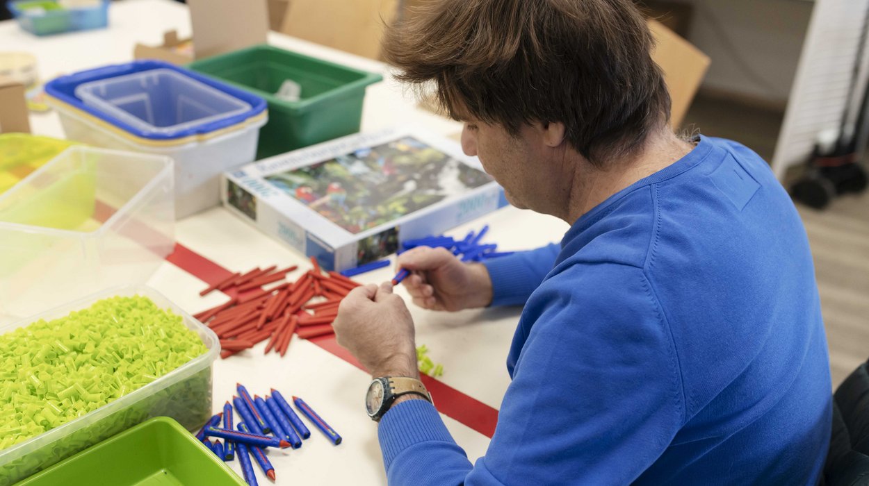 AB_Graz-West_Jugend-am-Werk-Steiermark Ein Mann mit Behinderung sitzt an einem Tisch und baut Kreidestifte in unterschiedlichen Farben zusammen