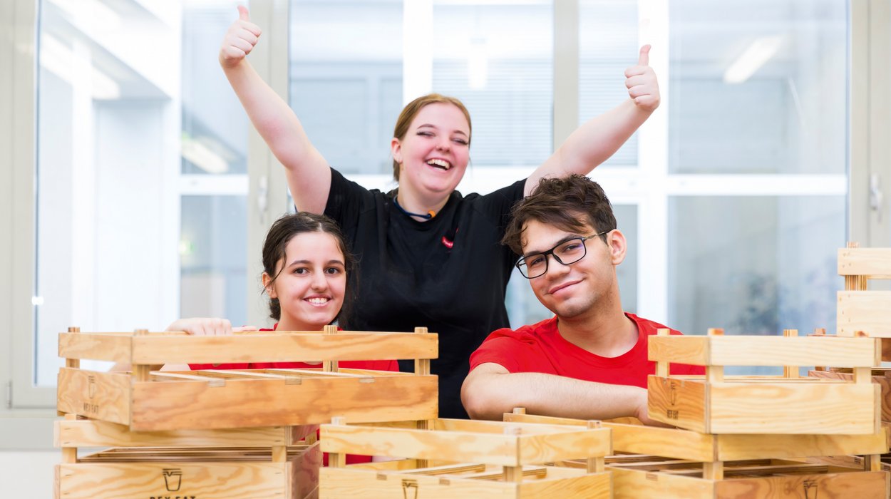 Produktionsschule_Jugend-am-Werk-Steiermark Drei Jugendliche glücklich hinter einem Stapel Holzkisten, die sie selbst gebaut haben