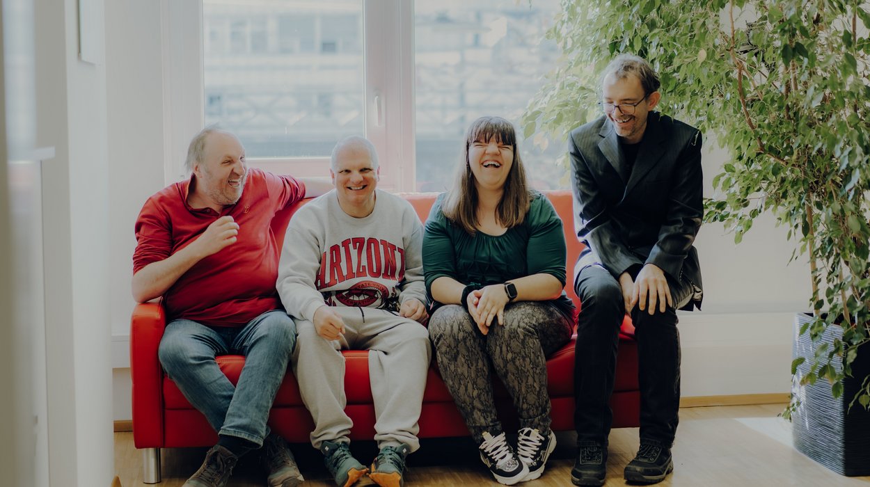 Interessensvertretung_Jugend-am-Werk-Steiermark Vier Menschen mit Behinderung sitzen vor einem großen Fenster auf einer Couch, alle lachen