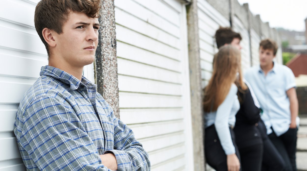 ein Jugendlicher mit einem nachdenklichen Blick und verschränkten Armen steht an einer Wand, im Hintergrund zwei Meter weiter unterhalten sich drei weitere Jugendliche